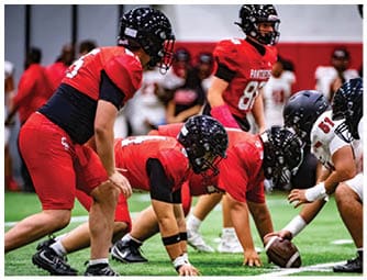 Several people in red and white football uniforms and helmets are positioned on a green field preparing for a play during a game. Description generated by AI