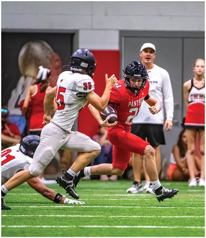 A person in a red football uniform is running with the ball while two people in white uniforms attempt to tackle them on an indoor field with spectators and cheerleaders in the background. Description generated by AI