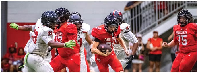 People wearing football helmets and uniforms are actively engaged in a play on an indoor field with spectators in the background. Description generated by AI