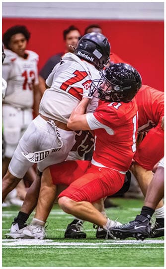 Two people in football uniforms are engaged in a tackle on an indoor field with others watching in the background. Description generated by AI