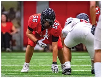 A person in a red football uniform is crouched in a ready position on a green field facing three people in white football uniforms preparing for a play. Description generated by AI