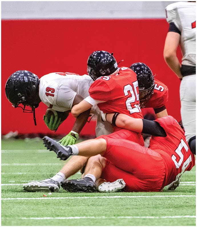 Several people in football uniforms are engaged in a tackle on an indoor field with a red wall background. Description generated by AI