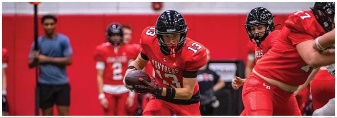 People wearing red football uniforms are actively playing on an indoor field with a person in the foreground holding a football and others preparing to engage. Description generated by AI