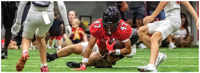 A person wearing a red football uniform with the number 47 is running with the ball while being tackled by a person in a white uniform on a green field with other players nearby. Description generated by AI