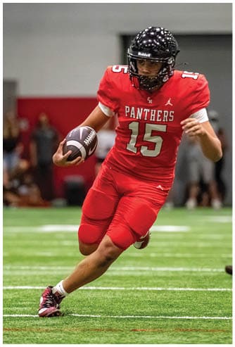 A person wearing a red football uniform with the word Panthers and number 15 is running on a green indoor field while holding a football. Description generated by AI