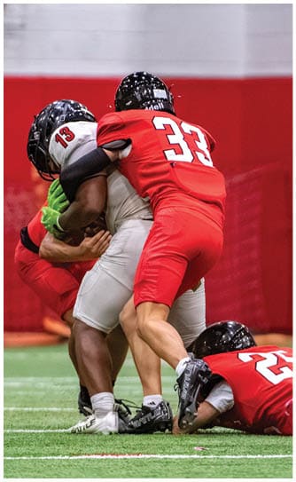 Three football players in red uniforms tackle a player in a white uniform on an indoor field while another player in red watches nearby. Description generated by AI