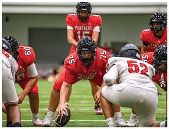A group of football players in red and white uniforms are positioned on a green indoor field preparing for a play. Description generated by AI