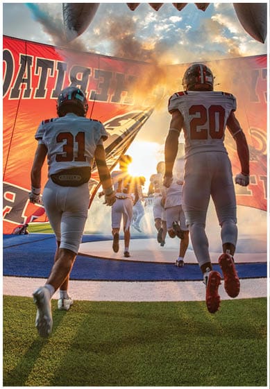 People in football uniforms run onto a field through a large red banner with sunlight and smoke creating a dramatic entrance. Description generated by AI