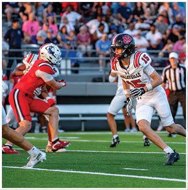 A person in a white football uniform with the number 15 is running on a green field toward two people in red football uniforms while a referee and spectators watch from the stands. Description generated by AI