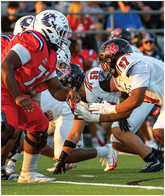 Several football players in helmets and uniforms are crouched and ready at the line of scrimmage on a green field during a game. Description generated by AI