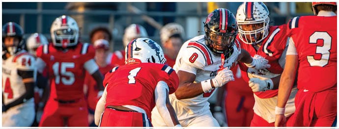Several people in football uniforms are engaged in a game on a field with one player in white preparing to make a move against players in red. Description generated by AI