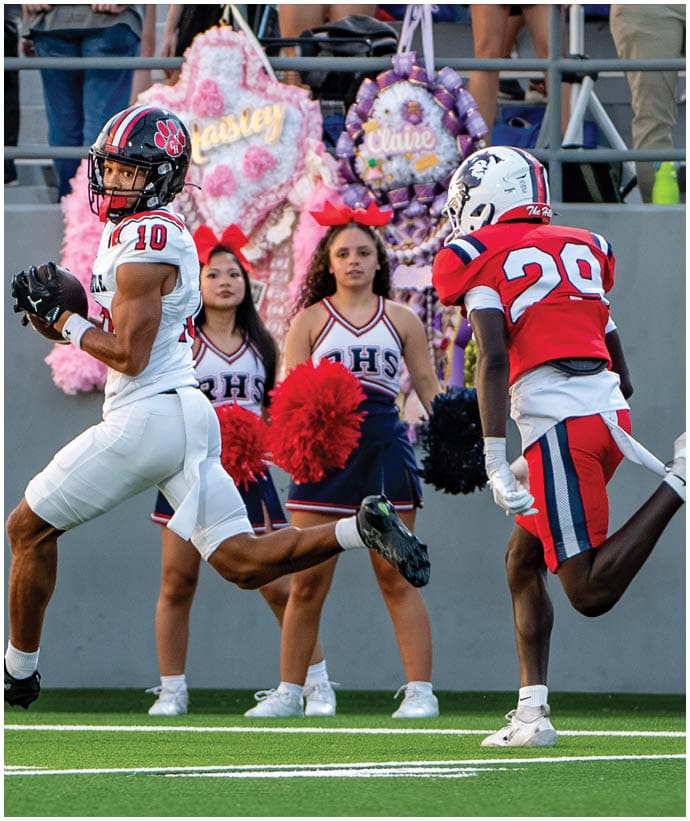 A person in a white football uniform catches a football while another person in a red football uniform approaches on a field with cheerleaders holding pom-poms standing in the background. Description generated by AI