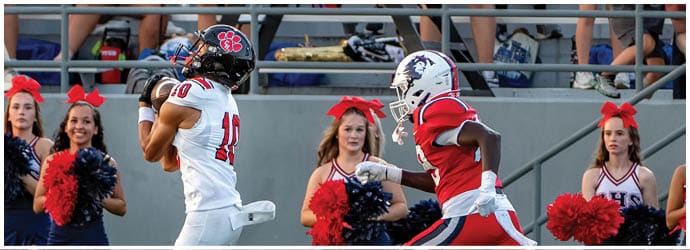 A person in a white football uniform catches a ball while another person in a red football uniform runs toward them with cheerleaders holding red and blue pom-poms standing in the background. Description generated by AI