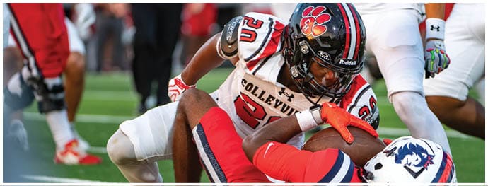 A person wearing a football uniform with a black helmet and white jersey is tackling another person in a red and blue football uniform on a grassy field. Description generated by AI