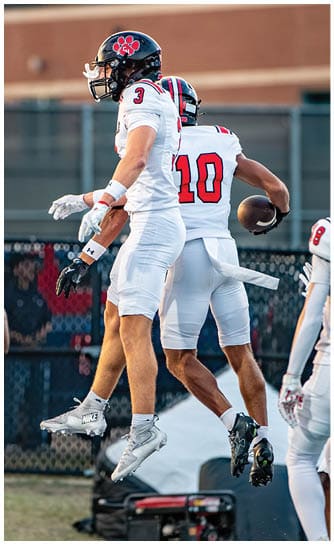 Two people wearing white football uniforms with red numbers are standing close together on a field. Description generated by AI