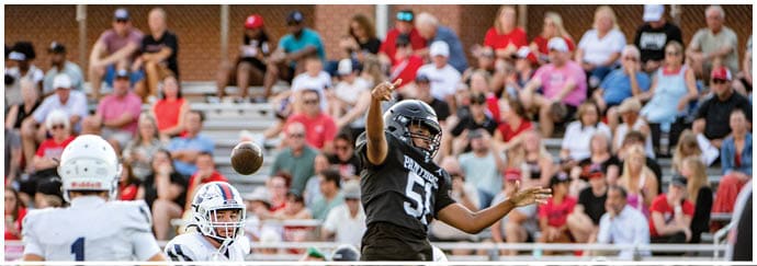 A person in a black football uniform is throwing a football while other players and a crowd of spectators watch from the stands. Description generated by AI
