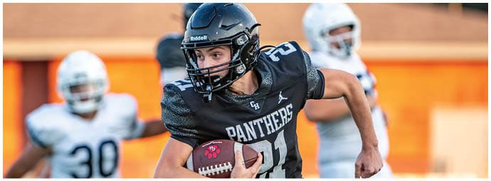 A person wearing a black football uniform with the word Panthers is running with a football while two people in white uniforms are in the background on a field. Description generated by AI