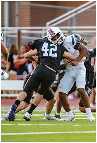 Two people in football uniforms are engaged in a tackle on a grassy field during a game. Description generated by AI