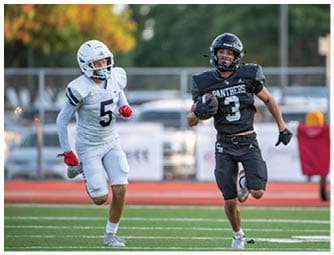 Two people in football uniforms are running on a field with one holding a football while the other chases. Description generated by AI