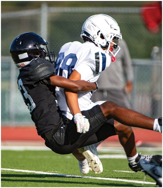 Two people wearing football helmets and uniforms are engaged in a tackle on a grassy field during a game. Description generated by AI