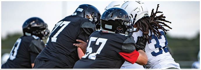 Three people in football gear are engaged in a play on a field, with two wearing black uniforms and one in a white uniform. Description generated by AI