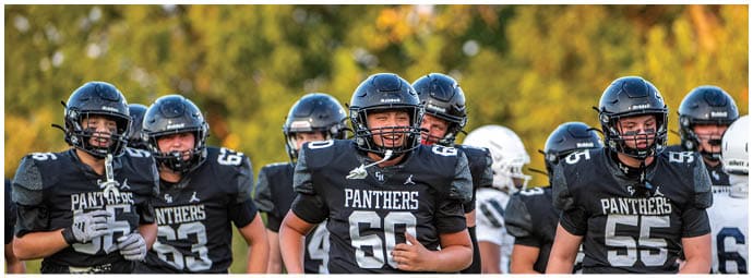 A group of people wearing black football uniforms with “Panthers" on the front are standing together outdoors on a field with trees in the background. Description generated by AI