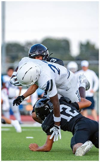 Two people in football uniforms are engaged in a tackle on a grassy field during a game. Description generated by AI