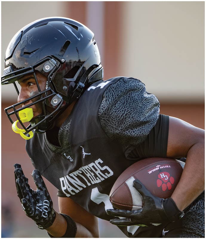 A person wearing a black football helmet and uniform with “PANTHERS" on the jersey is running while holding a football. Description generated by AI