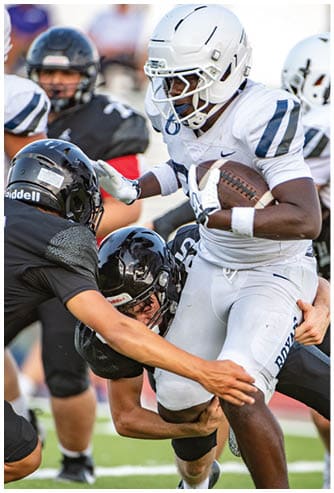 A person in a white football uniform is running with the ball while two people in black uniforms attempt to tackle them on a football field. Description generated by AI
