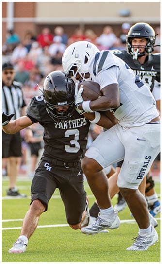 A person in a black Panthers football uniform is attempting to tackle another person in a white football uniform who is holding the ball tightly during a game. Description generated by AI