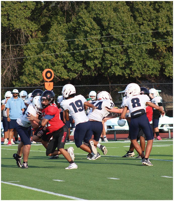People wearing football helmets and uniforms are engaged in a play on a field with a chain marker and trees in the background. Description generated by AI