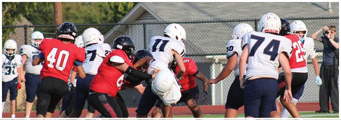 A group of people wearing football uniforms in red and white are engaged in a play on a football field with a person in the background taking a photo. Description generated by AI