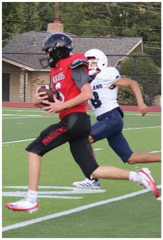 Two people in sports uniforms are running on a football field with one person holding a football while the other chases behind. Description generated by AI