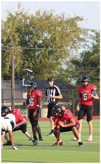 People in football uniforms are positioned on a field preparing for a play with a referee standing behind them and a coach in a red shirt observing from the side. Description generated by AI