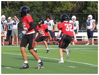 Three people in red football uniforms are on a green field with several people in white and blue uniforms standing near a fence in the background. Description generated by AI