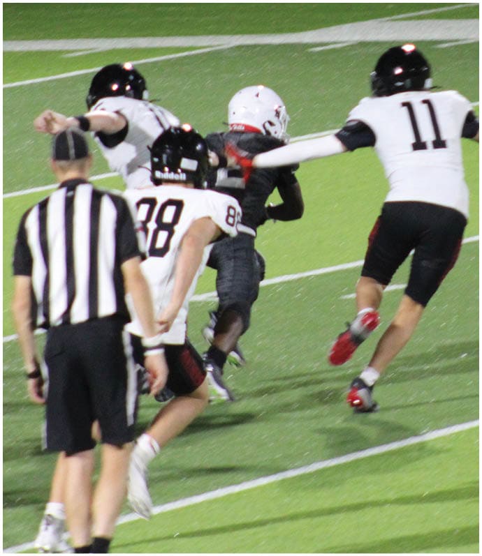 People wearing football helmets and uniforms are engaged in a play on a field with a chain marker and trees in the background. Description generated by AI