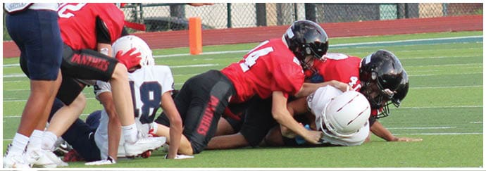 A group of people wearing football uniforms in red and white are engaged in a play on a football field with a person in the background taking a photo. Description generated by AI