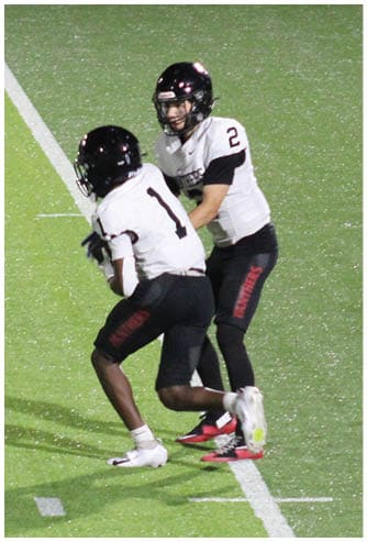Two people in sports uniforms are running on a football field with one person holding a football while the other chases behind. Description generated by AI