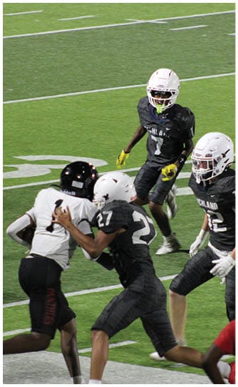 People in football uniforms are positioned on a field preparing for a play with a referee standing behind them and a coach in a red shirt observing from the side. Description generated by AI