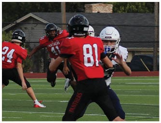 Three people in red football uniforms are on a green field with several people in white and blue uniforms standing near a fence in the background. Description generated by AI