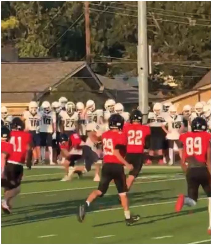 People wearing football helmets and uniforms are engaged in a play on a field with a chain marker and trees in the background. Description generated by AI