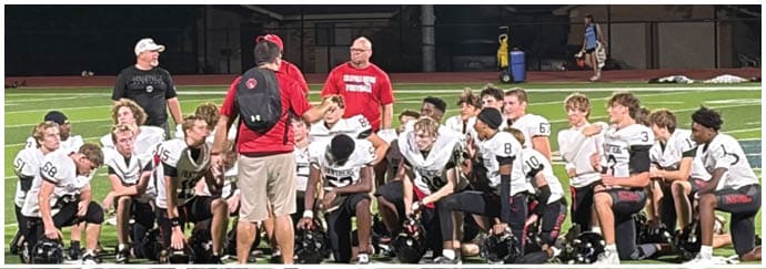 A group of people wearing football uniforms in red and white are engaged in a play on a football field with a person in the background taking a photo. Description generated by AI