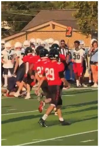 Two people in sports uniforms are running on a football field with one person holding a football while the other chases behind. Description generated by AI