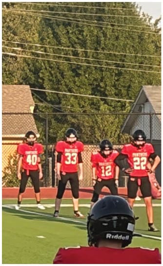 People in football uniforms are positioned on a field preparing for a play with a referee standing behind them and a coach in a red shirt observing from the side. Description generated by AI