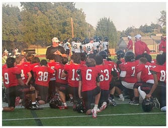 Three people in red football uniforms are on a green field with several people in white and blue uniforms standing near a fence in the background. Description generated by AI