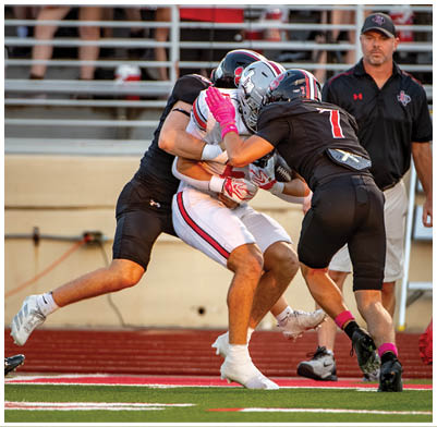 Two people in black football uniforms tackle a person in a white football uniform holding a football on a field with a coach and cheerleaders in the background. Description generated by AI
