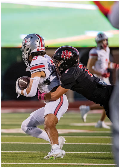 A person in a white football uniform is holding a football while being tackled by another person in a black football uniform on a field. Description generated by AI