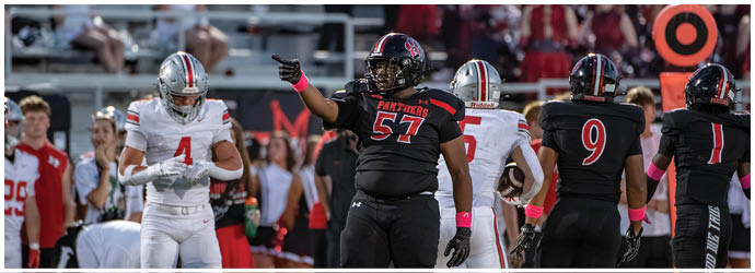 Players in black and red and white and silver football uniforms are on a field during a game with one player pointing and others standing nearby. Description generated by AI