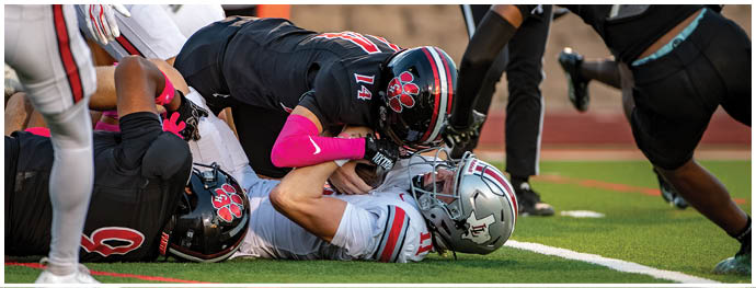 Two football players in black uniforms with pink accents are tackling a player in a white uniform during a game on a grassy field. Description generated by AI