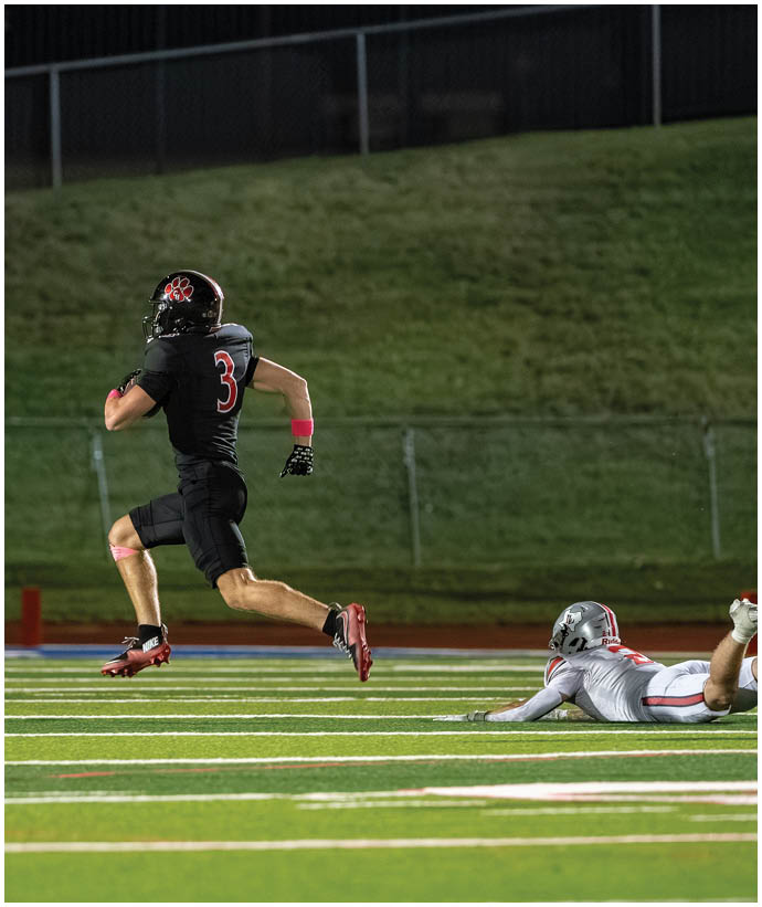A person in a black sports uniform with the number three is running on a football field while another person in a white uniform is lying on the ground behind them. Description generated by AI